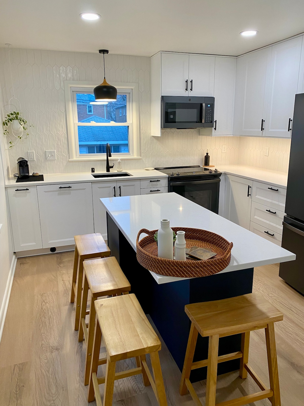 A modern kitchen is presented, featuring white cabinetry and a dark island. Four wooden stools are arranged around the island, which holds a decorative tray with bottles. Stainless steel appliances and a window provide ample light, illuminating a clean and functional space.