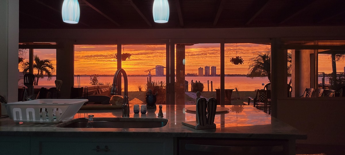A spacious kitchen area is visible, featuring a modern countertop and several kitchen accessories. Large windows showcase a stunning sunset over the water, illuminating the sky with vibrant oranges and yellows, while palm trees and distant buildings are framed outside.