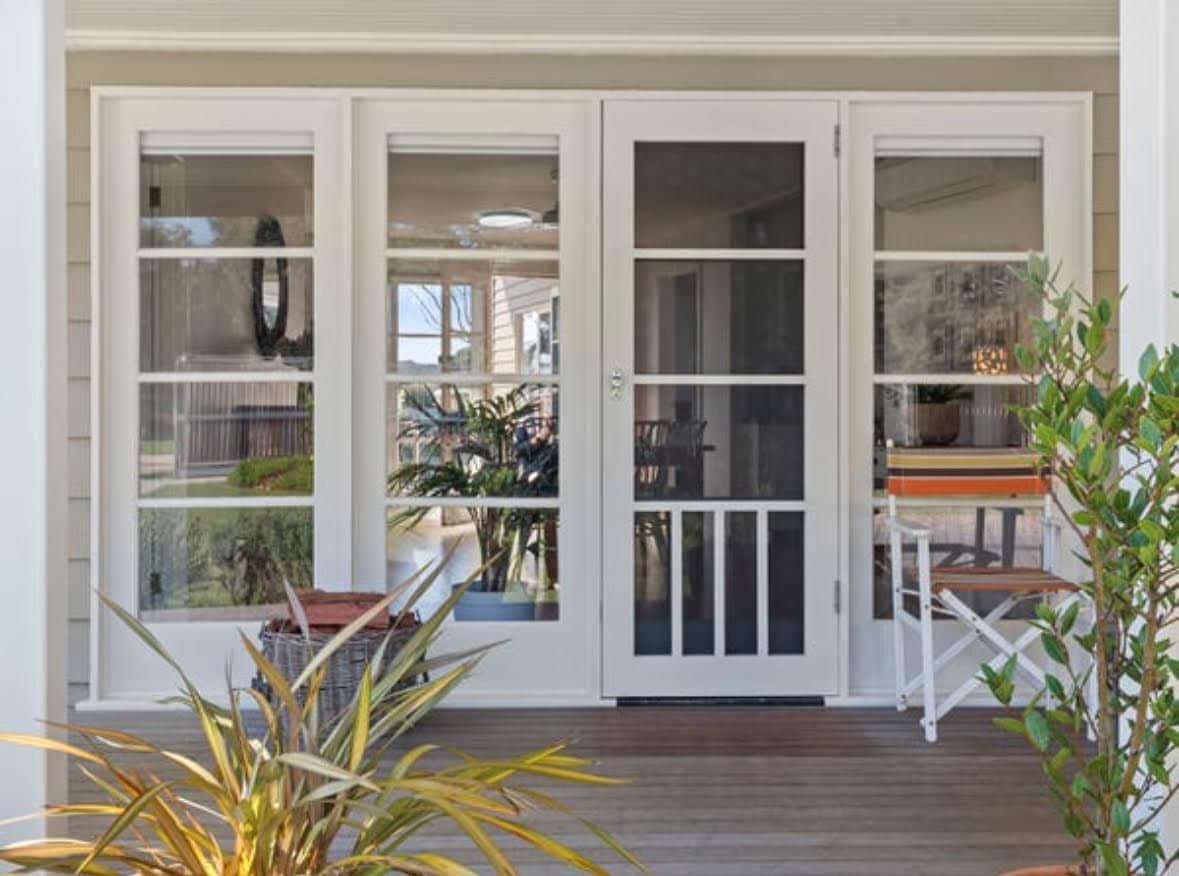 A screened porch entrance is presented, featuring white-framed glass doors. The space is bordered by potted plants, with a light wood floor enhancing the outdoor feel. Natural light filters through, inviting views of the surrounding garden area.