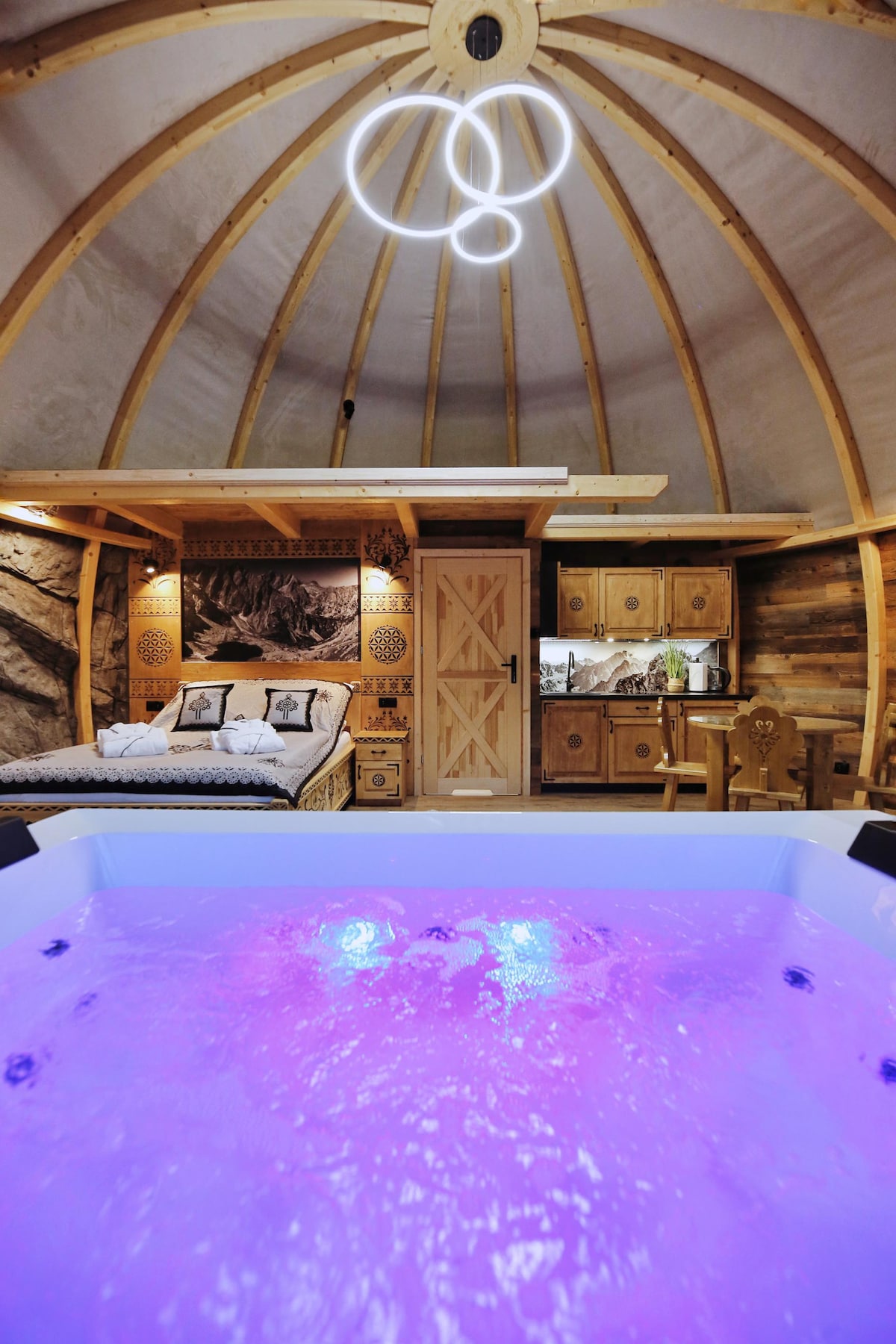 A unique interior space showcases a circular ceiling with wooden beams. A spacious bed is adorned with decorative pillows, and a well-equipped kitchenette is present in the background. A hot tub filled with water is illuminated in soft purple tones, adding a serene ambiance.