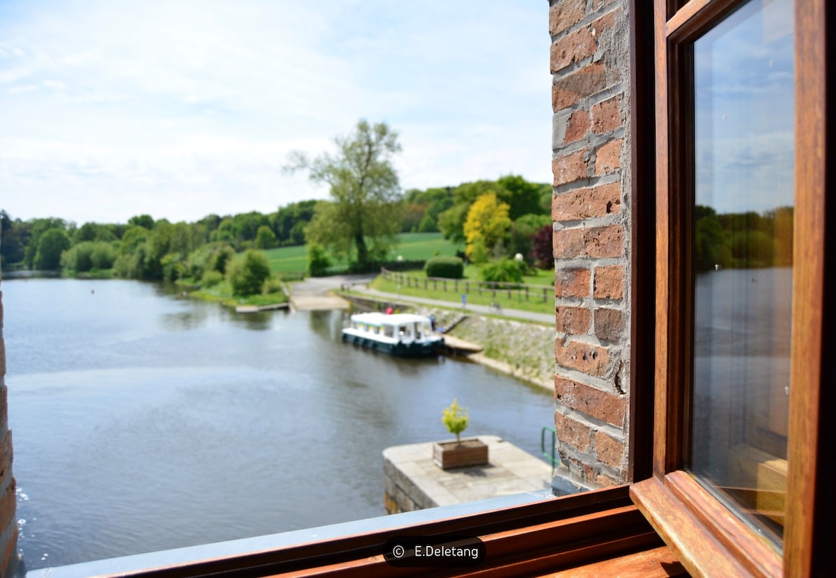 A serene view framed by a wooden window showcases the calm river and lush greenery along the banks. A small dock is visible with boats secured, and trees in various shades of green line the landscape. The atmosphere reflects the tranquility of the area.