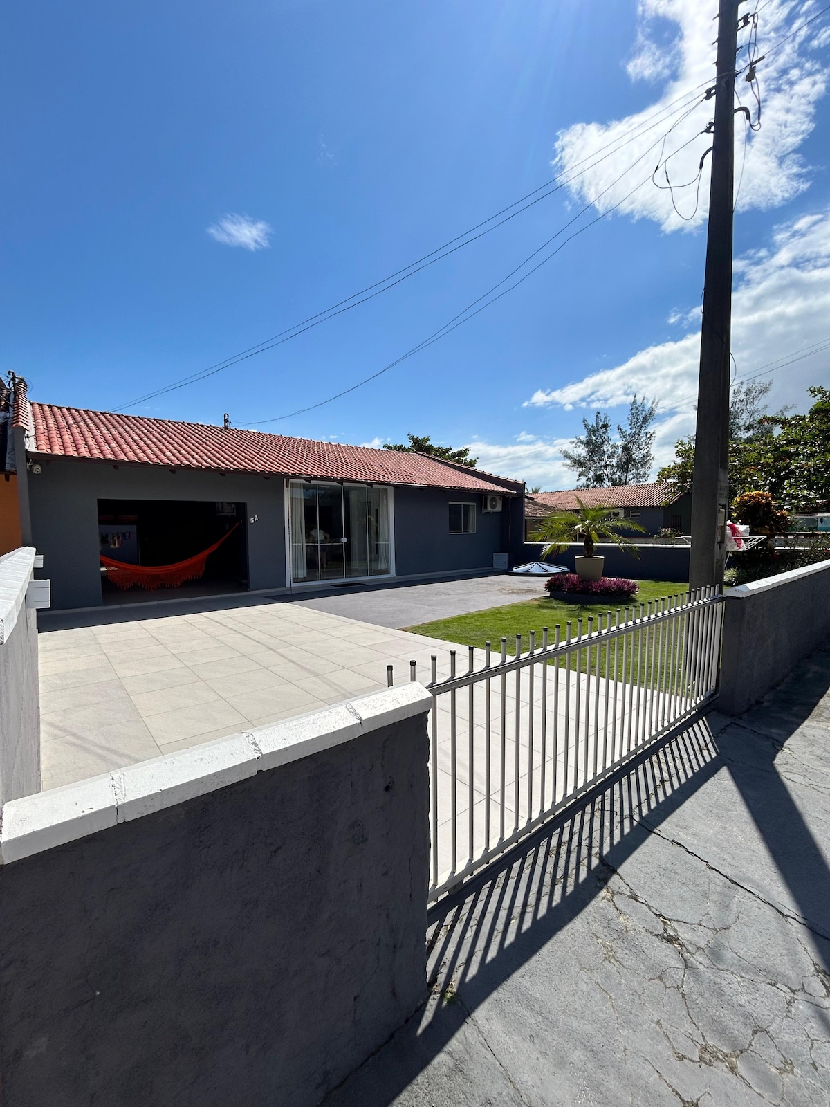 The exterior of the house features a well-maintained front yard with green grass and a paved area. Large glass doors allow natural light to enter, and a hammock is visible, creating a relaxed outdoor space. A low fence encloses the property.