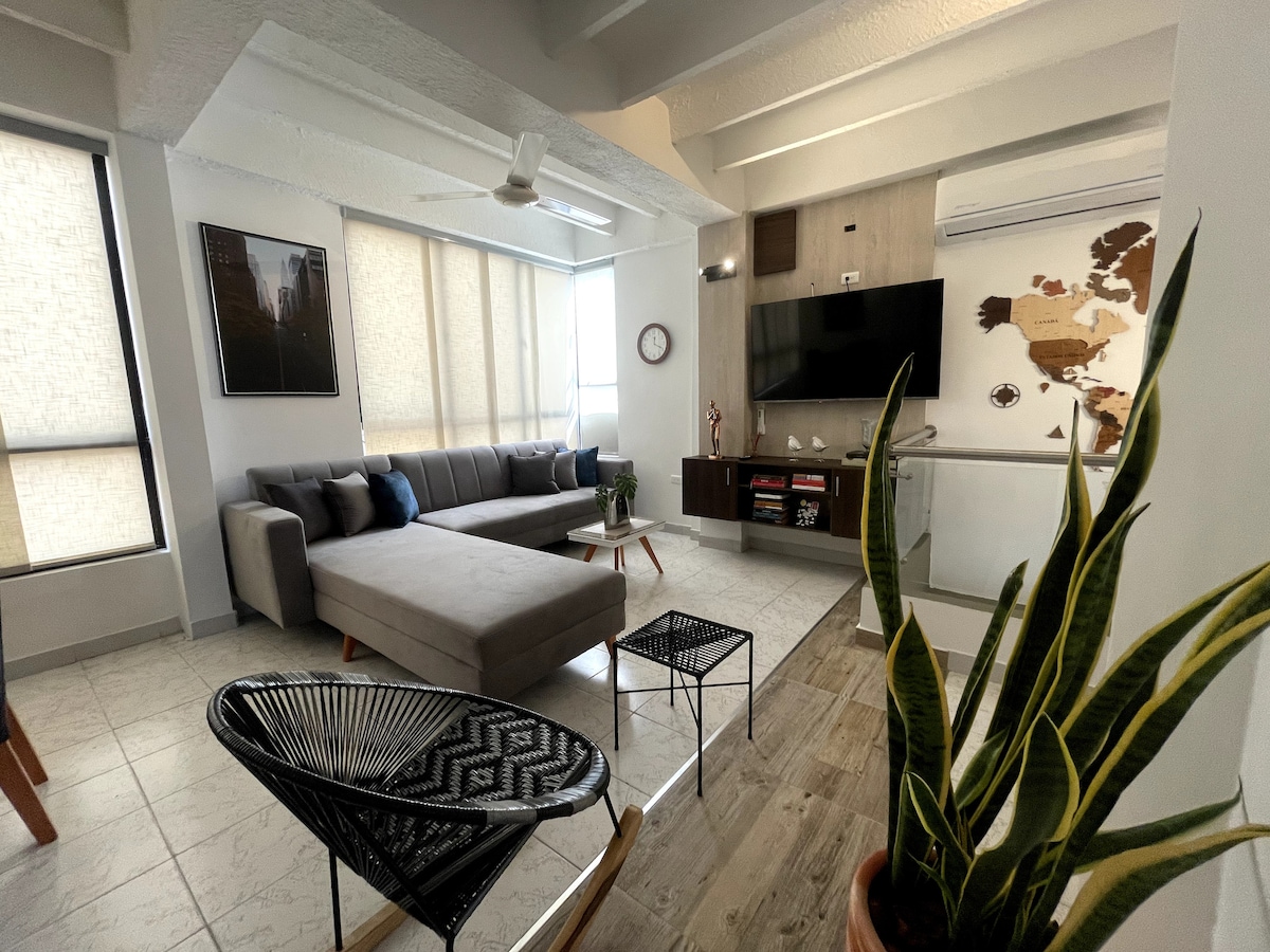 A modern living area is displayed with a spacious gray sectional sofa, a large television mounted on the wall, and a decorative map art piece. Natural light filters through the window shades, and a green plant adds a refreshing touch to the arrangement.