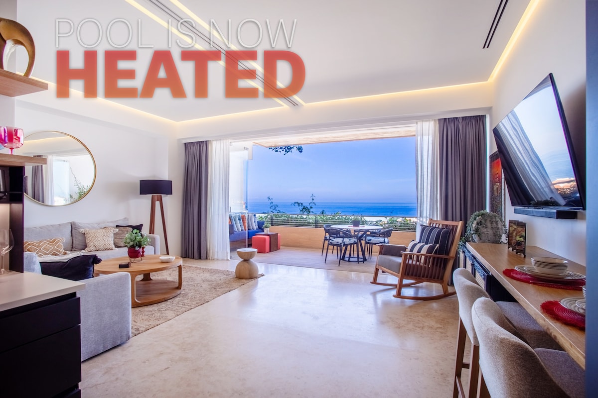 This modern living area features a comfortable sofa, stylish armchairs, and a wooden dining table. Large windows open to a view of Banderas Bay, allowing natural light to fill the space. Neutral tones and contemporary decor create a relaxed and inviting ambiance.