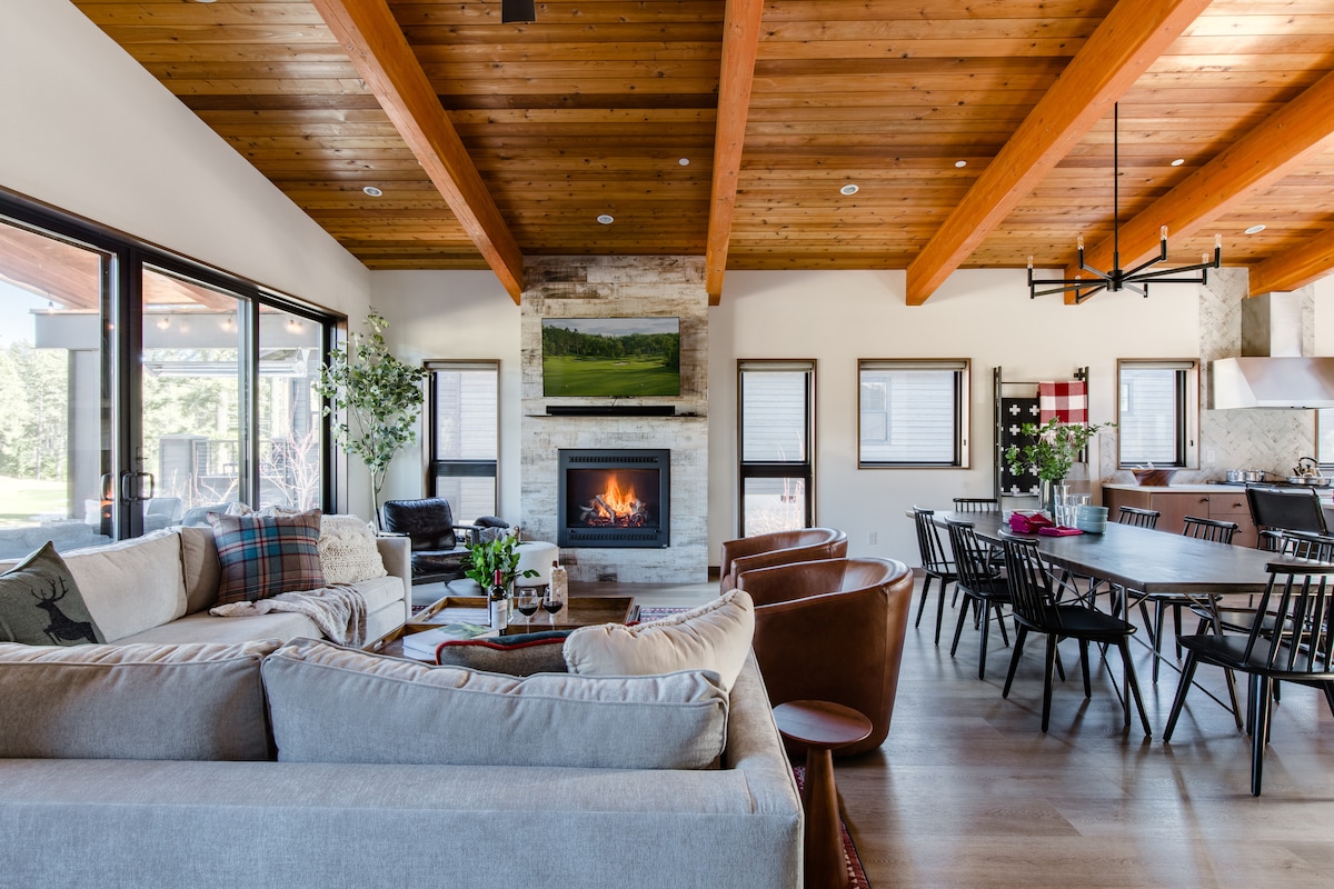 An open living area features a textured sectional sofa and comfortable leather chairs. A stone fireplace serves as a focal point under a wooden beam ceiling, while large windows provide natural light. A spacious dining table accommodates multiple guests, enhancing the communal atmosphere.