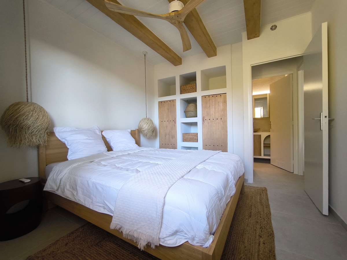 A spacious bedroom is featured with a wooden bed, topped with white linens. Two woven light fixtures hang above, complemented by light-colored walls and exposed timber beams. A doorway leads to a modern bathroom, and natural light brightens the room.