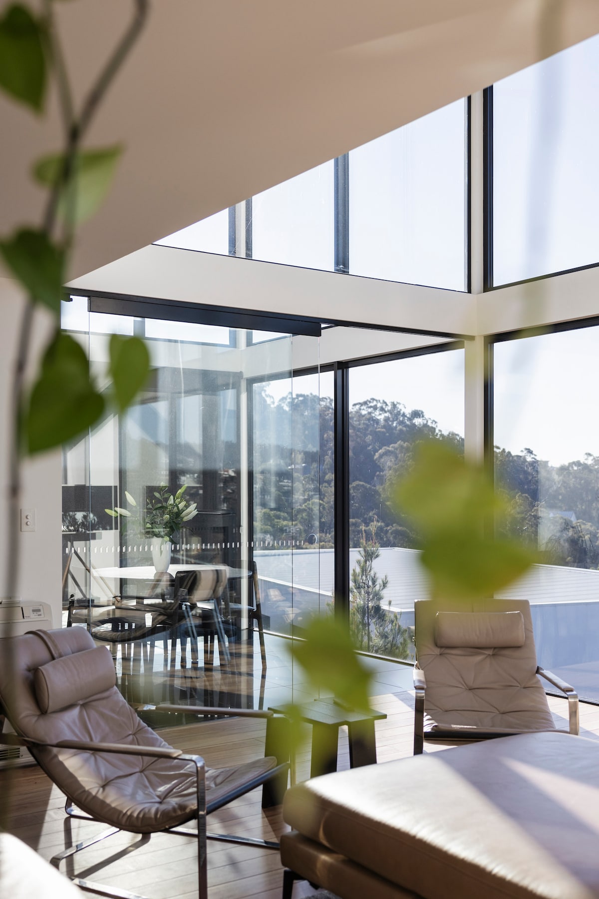 The living area features floor-to-ceiling windows that flood the space with natural light, offering stunning views of the surrounding landscape. Comfortable seating is arranged nearby, complemented by greenery that adds a touch of nature to the modern architecture.