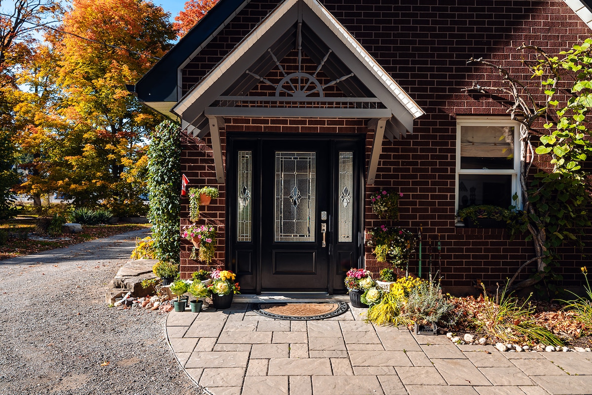 The entrance showcases a well-maintained brick façade, featuring a decorative front door with glass inserts. Surrounding flowers in various pots add vibrant colors, complemented by seasonal foliage from nearby trees. The stone pathway leads to the door, creating an inviting access point.