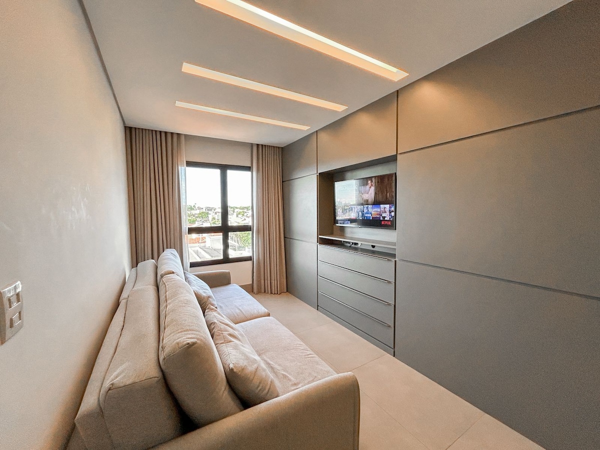 A modern living area is highlighted by a light-toned, spacious sofa and an entertainment unit with a flat-screen TV. Large windows allow ample natural light, offering a view of the city. Soft, neutral curtains complement the minimalist design.