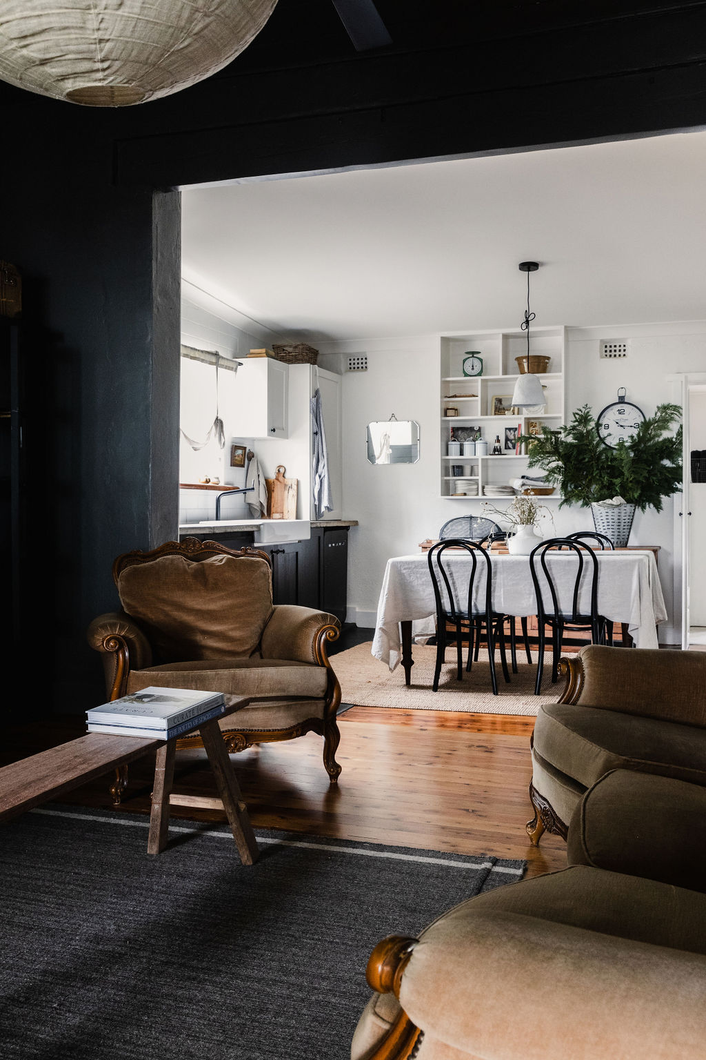 A cozy dining area features a white table set with dark chairs, alongside a well-organized kitchen. Dark walls contrast with light cabinetry, while potted plants add a touch of greenery. Vintage armchairs are positioned invitingly in the foreground, enhancing the warm atmosphere.