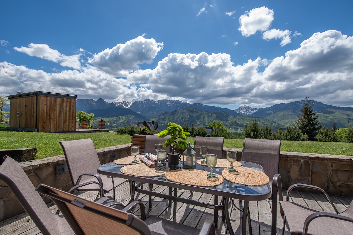 A spacious terrace is presented with a large dining table surrounded by six chairs, all set against a stunning mountain backdrop. Soft clouds drift across the blue sky, enhancing the view of the surrounding landscape. A wooden hot tub is located in the background.