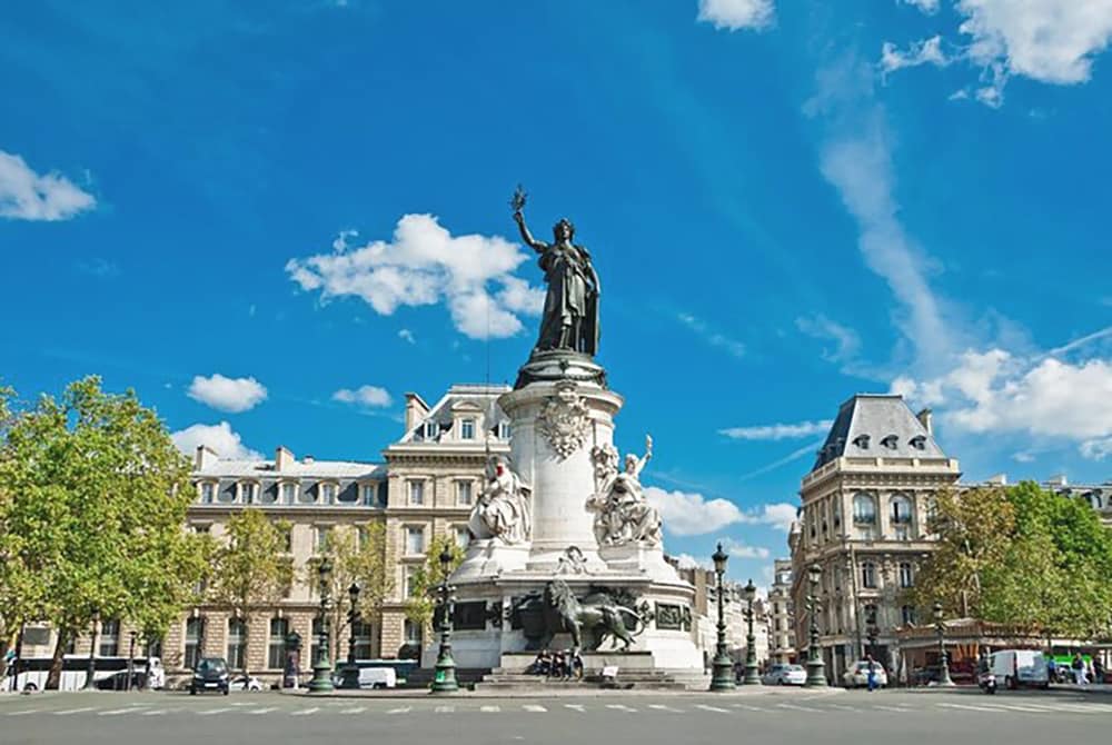 A large monument stands prominently in the center of a spacious square, surrounded by trees and classical buildings. The sky is bright blue with fluffy white clouds, enhancing the open atmosphere. Statues depict historical figures, while the scene conveys a sense of calm in the heart of the city.