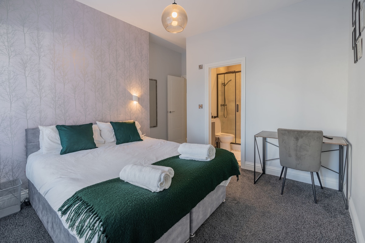A spacious bedroom features two single beds arranged together, adorned with white bedding and dark green throw blankets. A small desk with a grey chair is positioned to one side, while natural light illuminates the room from a nearby window, complemented by subtle wall patterns.