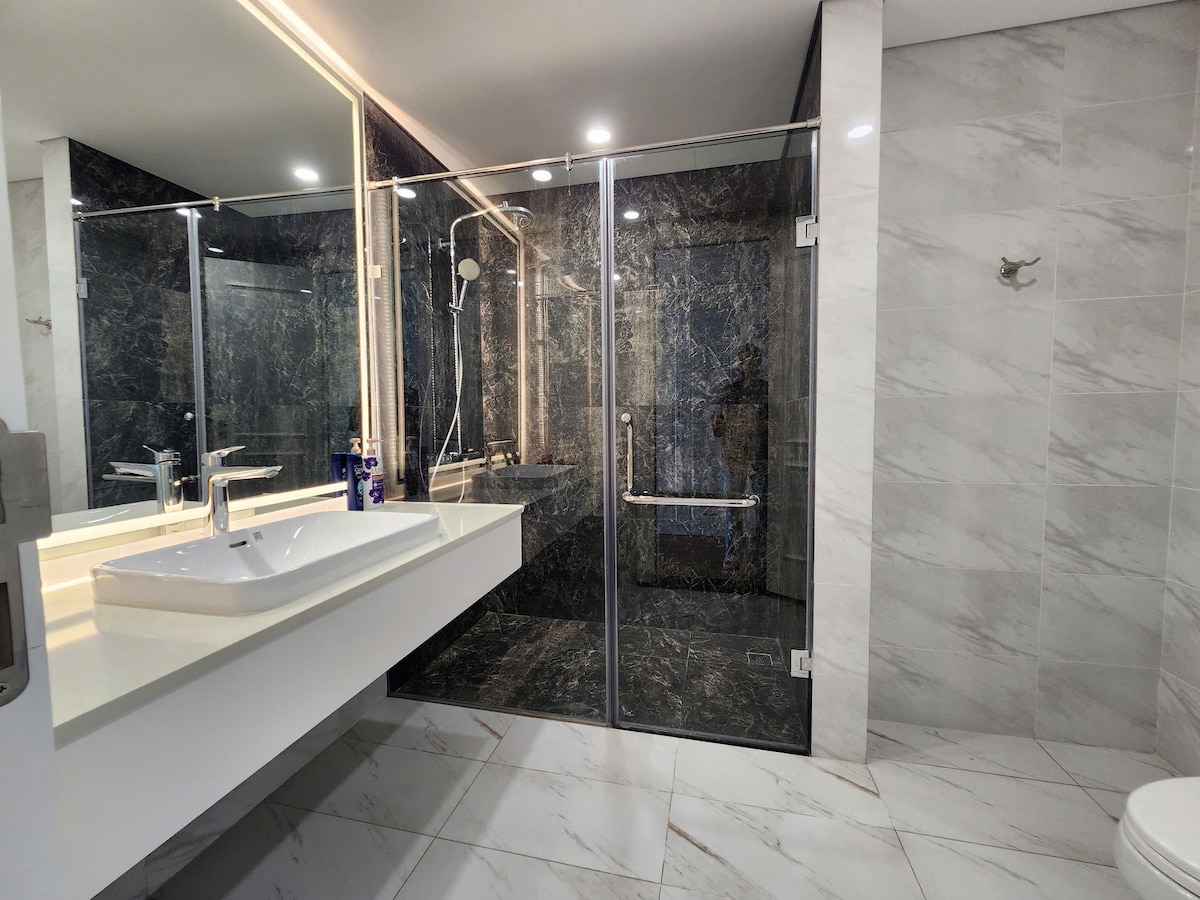 The spacious bathroom features sleek marble tiles and a modern design. A glass-enclosed shower is visible, alongside a large sink with contemporary fixtures. Soft lighting enhances the elegant atmosphere, and the layout provides a clean, open feel.