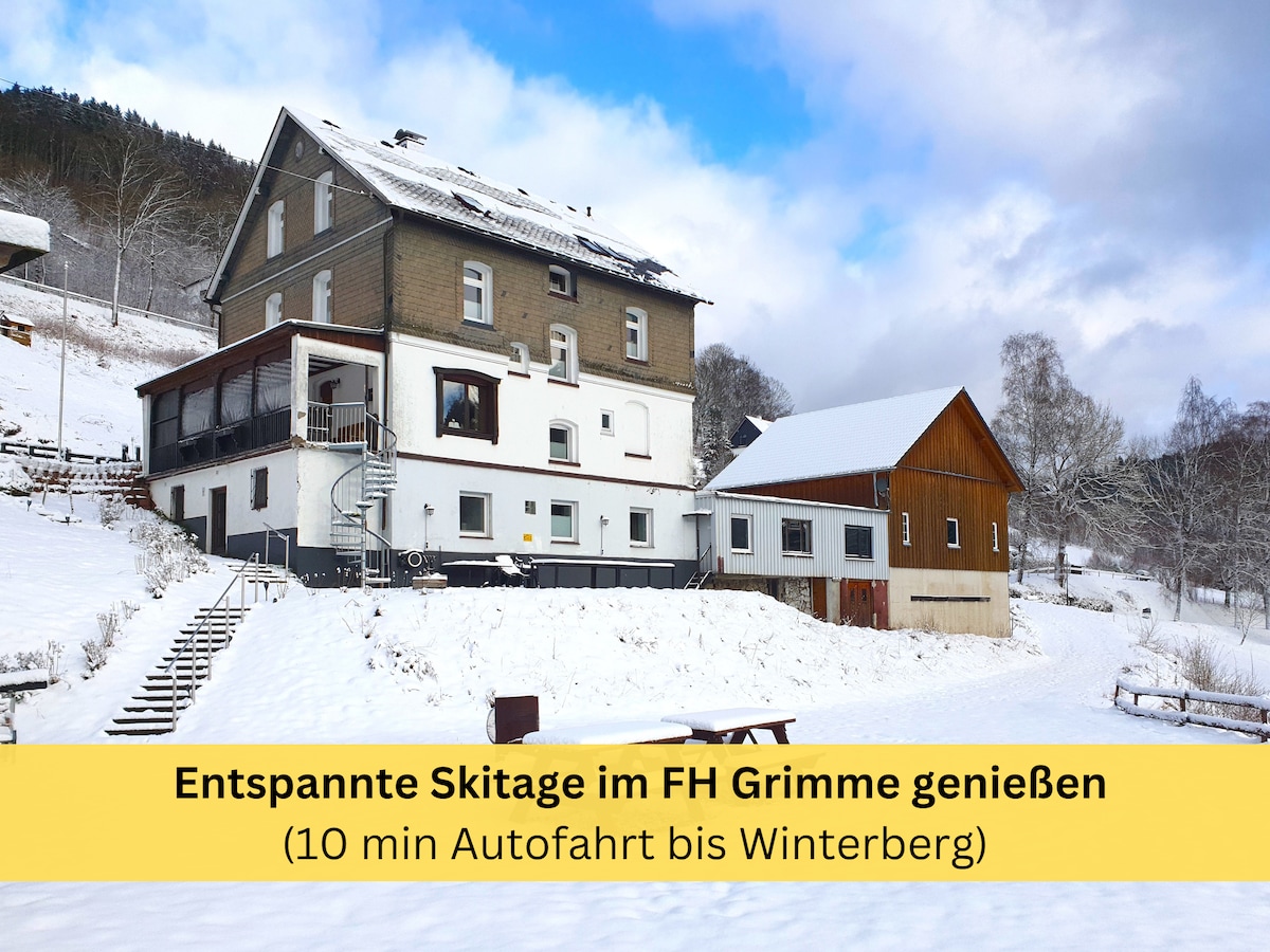 The exterior of the FH Grimme is set against a snowy landscape, showcasing a three-story building with a wooden annex. A staircase leads to the main entrance, while a view of the surrounding hills and trees emphasizes the serene winter atmosphere.