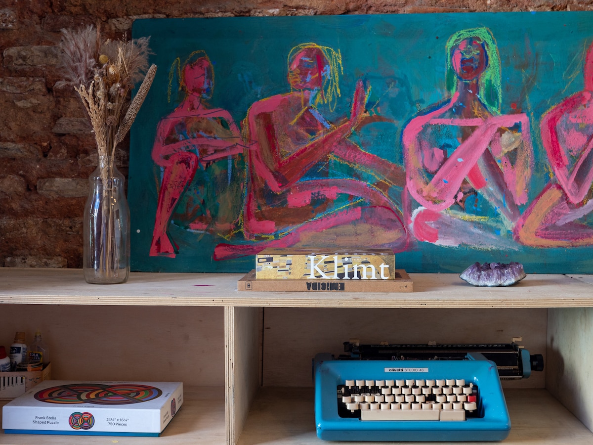 A vibrant artwork inspired by Klimt is displayed against a textured wall, featuring abstract representations of figures in bright colors. Below, a vintage typewriter in blue resides on a wooden shelf, accompanied by decorative items including a vase with dried flowers and an art supply box.