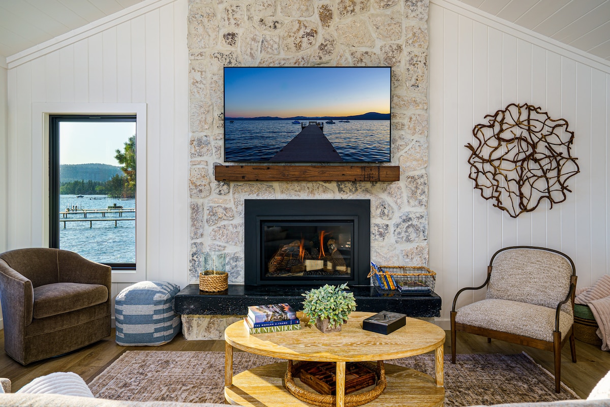 An inviting living area features a stone fireplace with a mounted television above, displaying a serene lakeside view. Comfortable seating options include two armchairs and a patterned lounge chair. Natural light filters through a window, highlighting a coffee table with decor and books.