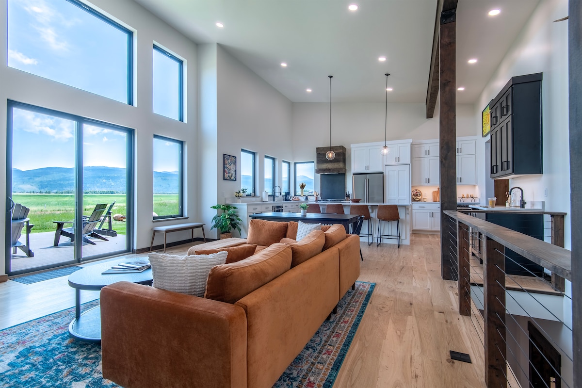 A spacious living area is filled with natural light from large windows, which offer views of the surrounding landscape. A comfortable sofa faces a coffee table, complemented by a decorative area rug. The open layout includes a modern kitchen visible in the background.