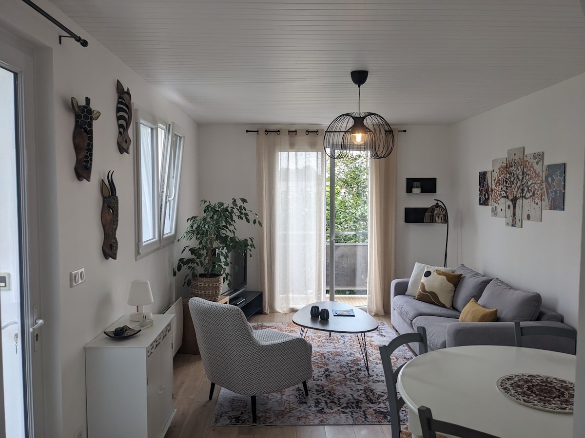 A cozy living space is highlighted by natural light filtering through sheer curtains. A comfortable sofa, accent chair, and dining table contribute to a welcoming atmosphere. Decorative wall art and greenery add visual interest, enhancing the room’s relaxed ambiance.