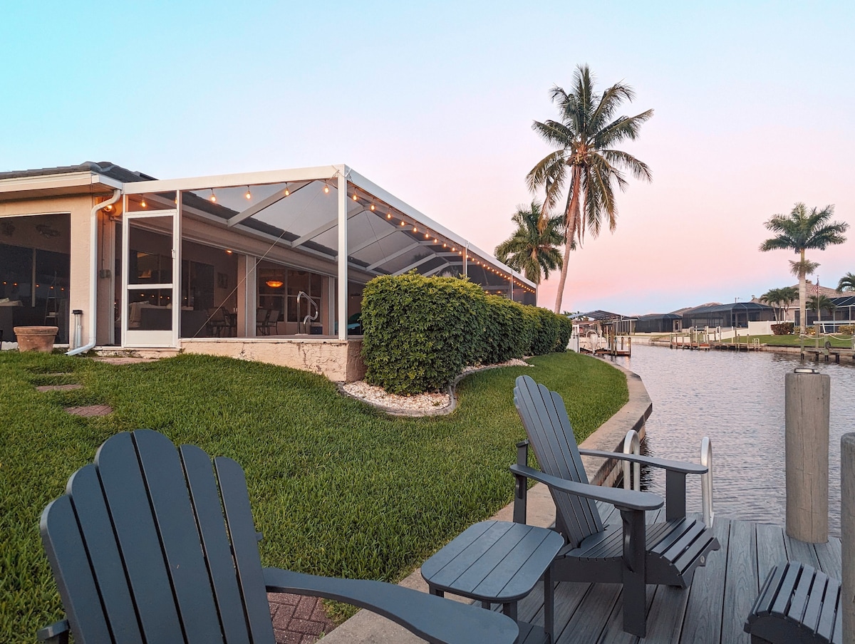 A serene waterfront view is presented, featuring a neatly landscaped area with lush greenery and palm trees. Comfortable adirondack chairs are positioned along the dock, offering a place to relax while enjoying the peaceful canal setting. The screened lanai is visible in the background.