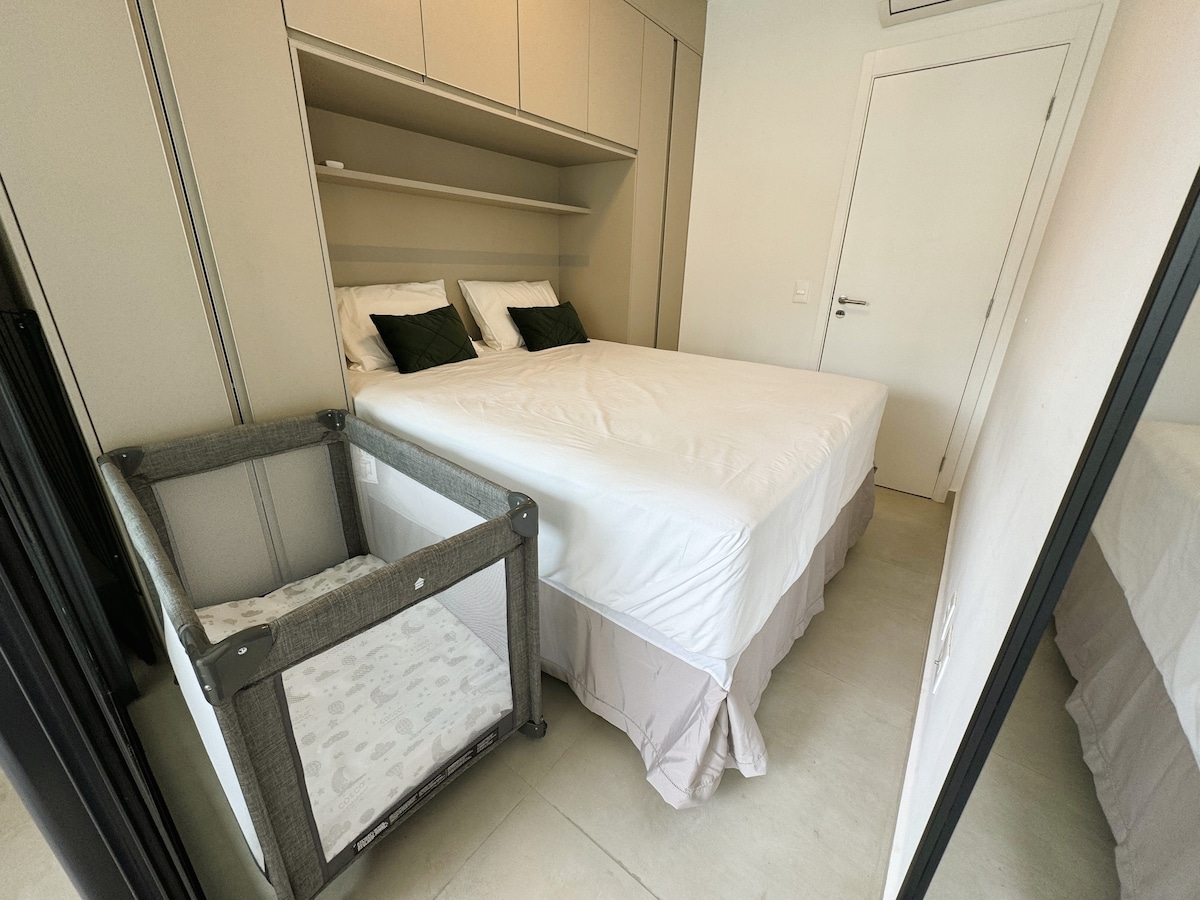 The bedroom features a spacious bed made with fresh linens, accompanied by a portable crib for infants. Soft green accents on decorative pillows complement the neutral color palette. Built-in shelves provide storage above the bed, and the room is illuminated by natural light.