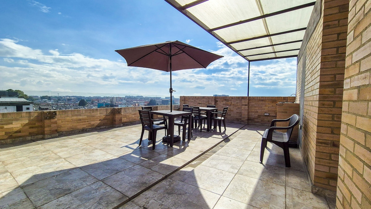 A spacious terrace is presented, featuring a large table surrounded by six chairs. An umbrella offers shade from the sun, and the area is paved with tiles, providing a smooth surface. The view includes distant cityscape and open sky, enhancing outdoor relaxation.