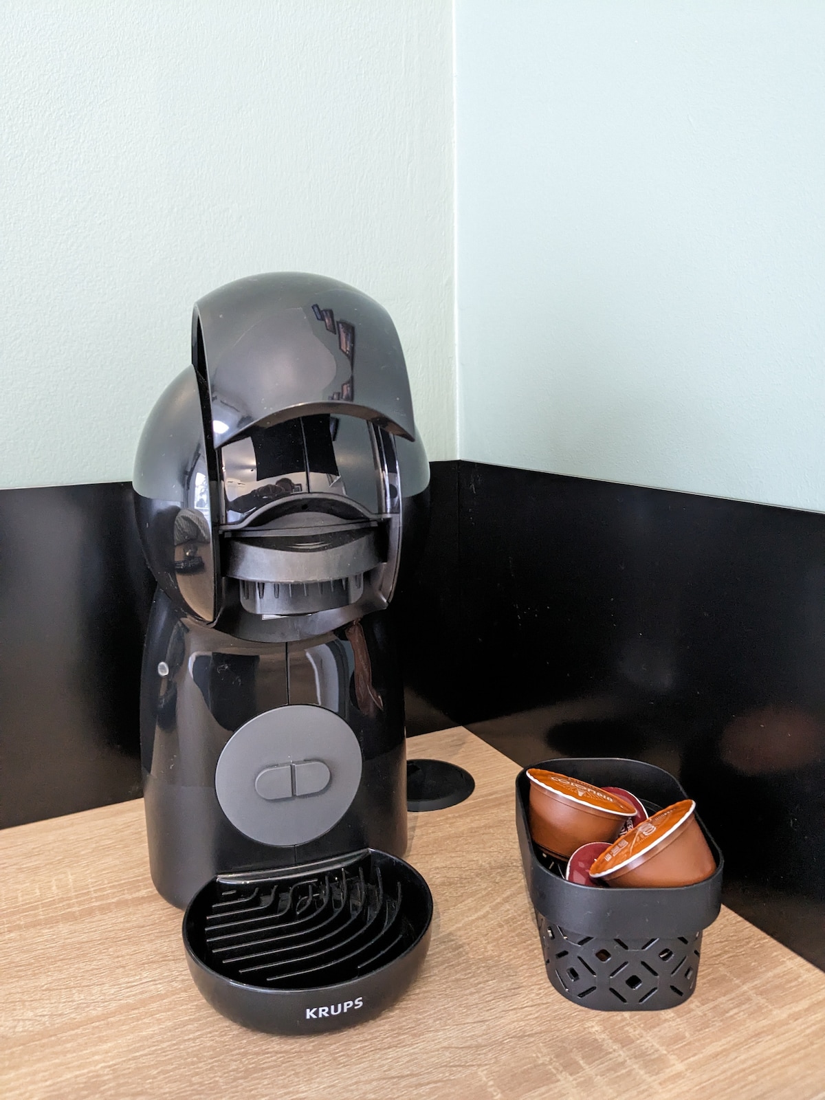 A sleek black coffee machine is positioned on a wooden countertop, accompanied by a small basket containing coffee pods. The machine features a simple button interface and a drip tray beneath, with a light green wall serving as a backdrop.