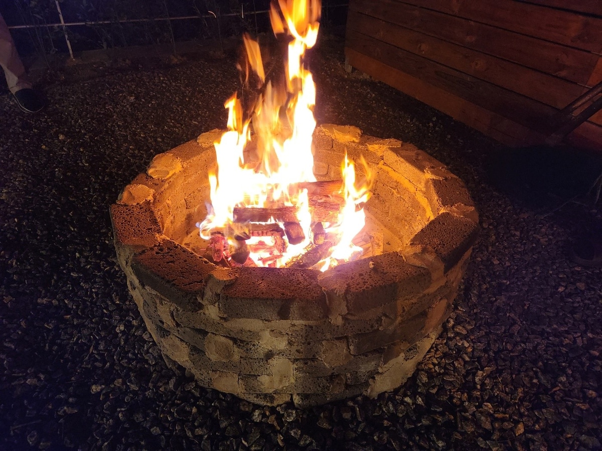 A circular stone fire pit is shown, filled with burning wood. Flames rise brightly from the center, casting a warm glow against the surrounding pebbled ground. The structure is made of layered stones, providing a rustic setting for outdoor gatherings.