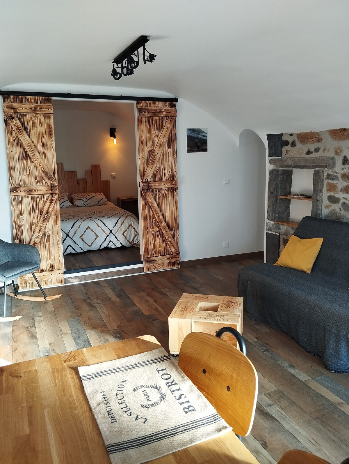 A cozy living area is visible, featuring a wooden dining table with chairs, and a gray sofa with a decorative yellow pillow. Sliding barn doors lead to the bedroom space, where a double bed with patterned bedding can be discerned. The room showcases natural wood flooring and rustic stone accents.