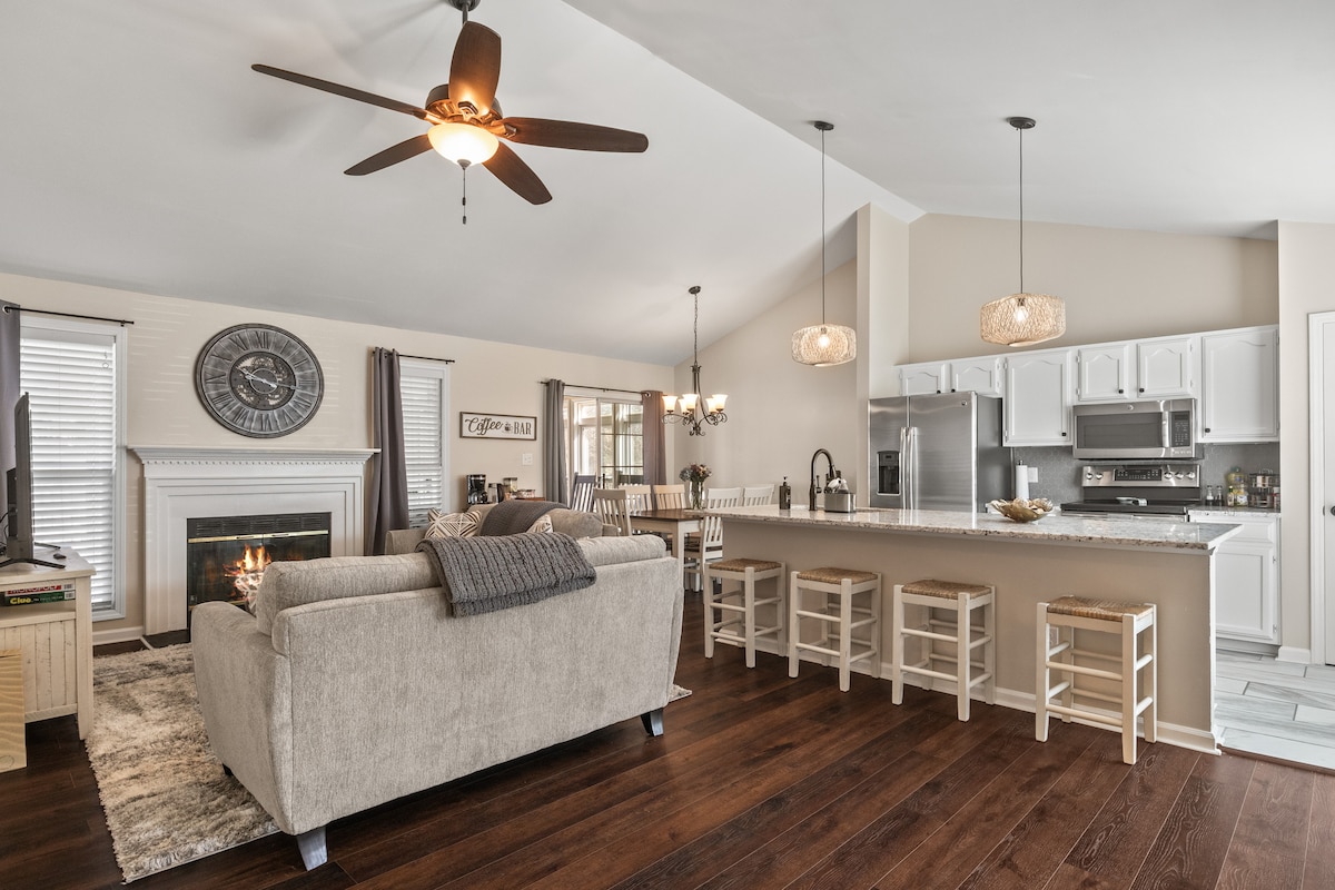 An open-concept living area features a light-colored sofa facing a fireplace. A large clock is mounted on the wall, and a dining area is visible nearby. The kitchen showcases modern appliances and bar seating, illuminated by stylish pendant lights.