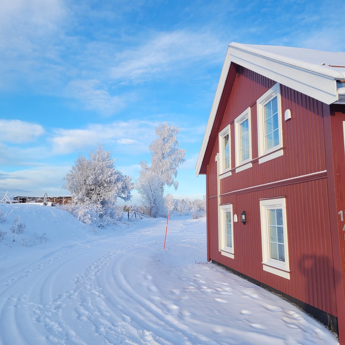 A red building with white trim is partially surrounded by a blanket of fresh snow. Tall trees covered in frost stand nearby, and a clear blue sky with scattered clouds provides a picturesque backdrop. A winding snowy path is visible leading towards the structure.
