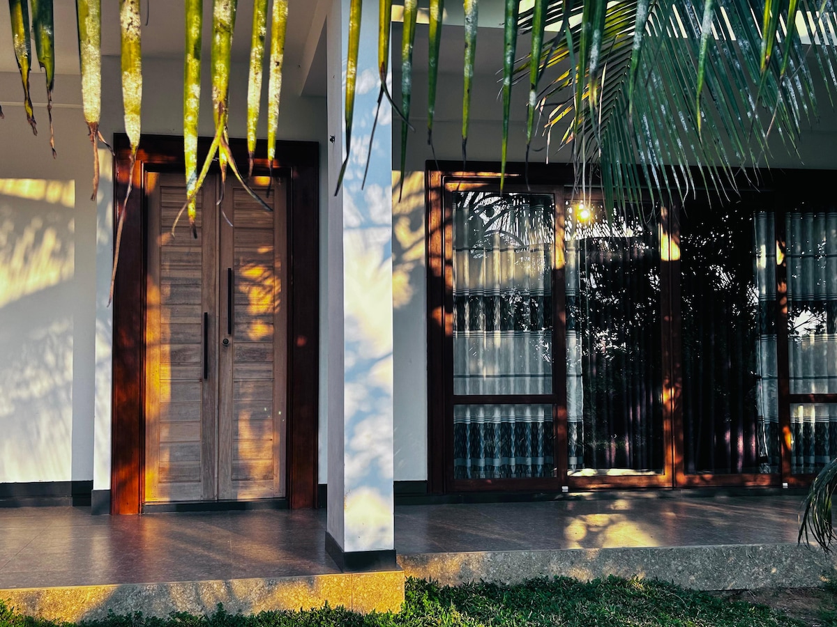 The entrance showcases beautifully crafted wooden doors, framed by lush green foliage. Natural light creates intricate shadow patterns on the walls, while large windows adorned with sheer curtains provide a glimpse into the interior space.