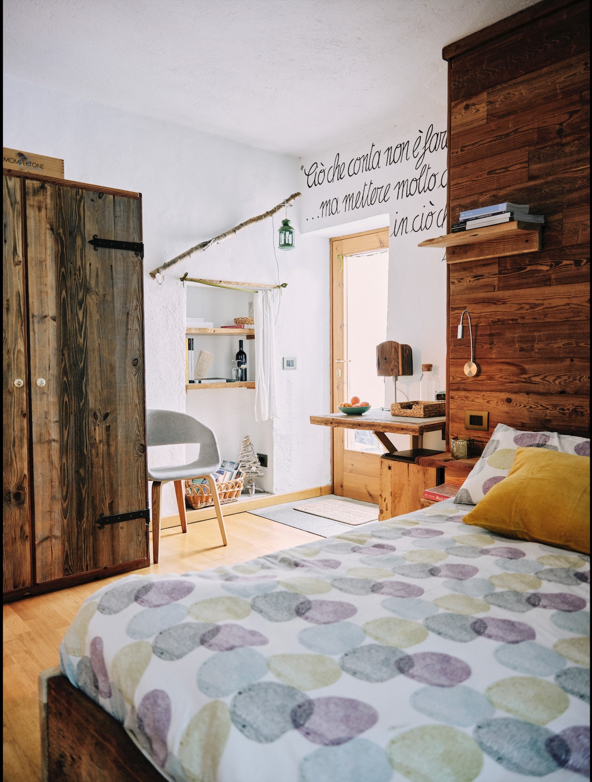 A cozy sleeping area is presented with a spacious bed, topped with a patterned duvet. Wooden accents on the walls complement a modern wardrobe. A small seating area includes a chair and table, while a door leads to an outdoor space, enhancing the natural light in the room.