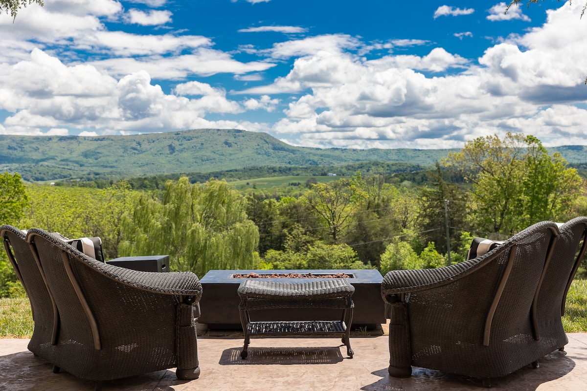 Seating is arranged around a central fire pit on a patio, offering views of rolling green hills and majestic mountains under a bright blue sky. Lush trees and a vibrant landscape are visible, enhancing the natural setting.