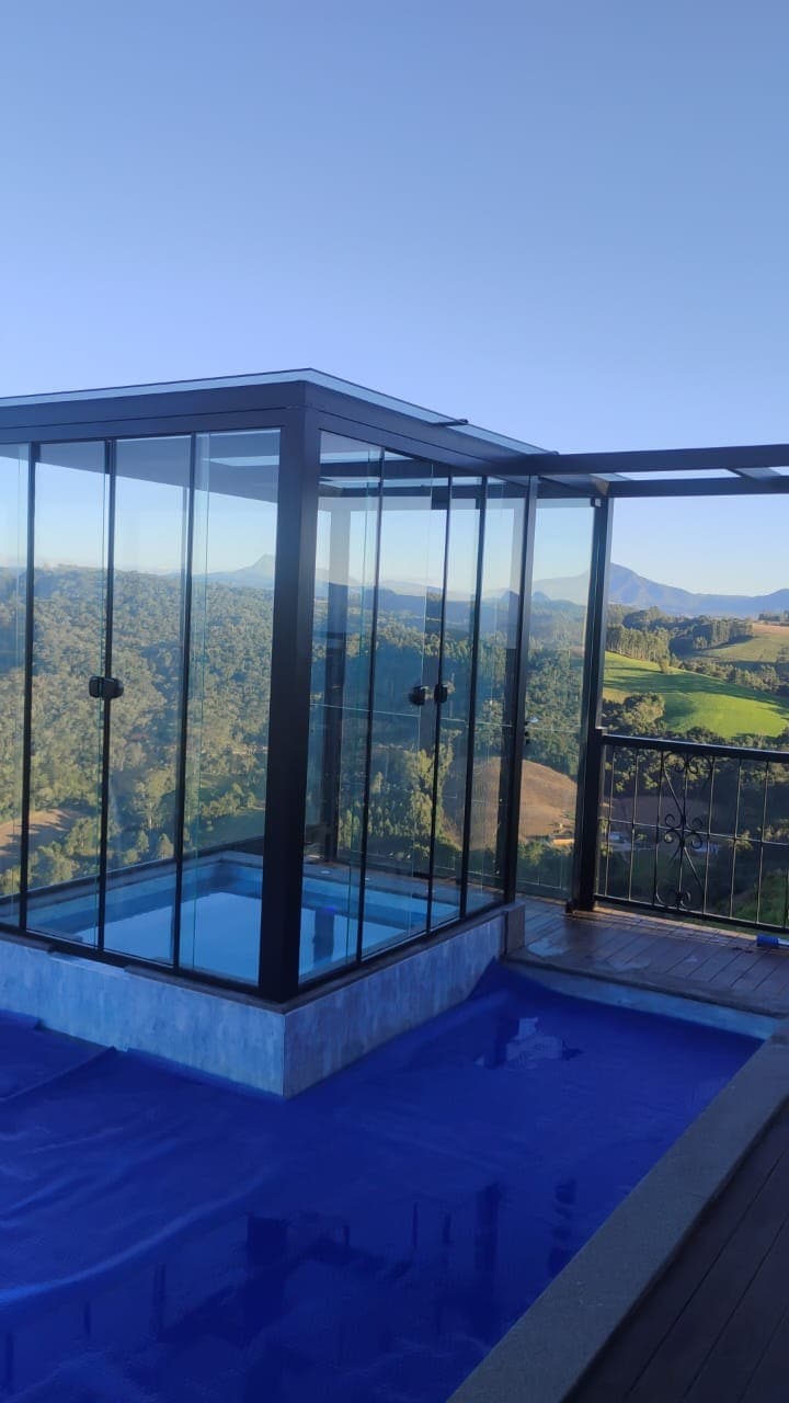 A glass-enclosed jacuzzi is positioned against a backdrop of rolling hills and a clear blue sky. The structure is outlined in sleek black framing, and the surrounding deck features a deep blue pool, enhancing the serene landscape view.