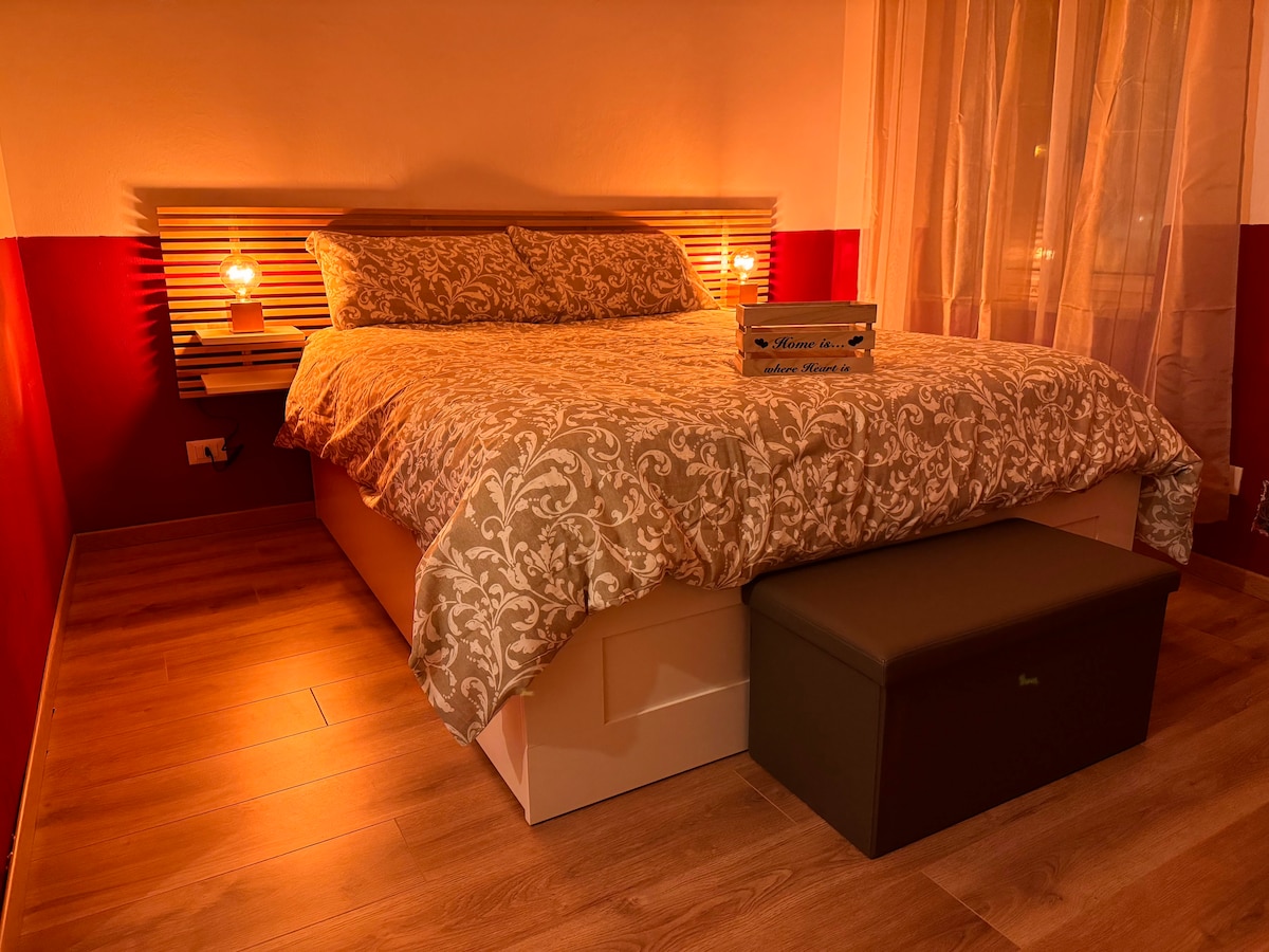 A well-lit bedroom features a spacious bed with decorative bedding. Soft bedside lamps provide warm illumination. A comfortable bench at the foot of the bed complements the warm color scheme, while sheer curtains allow natural light to filter through the window.