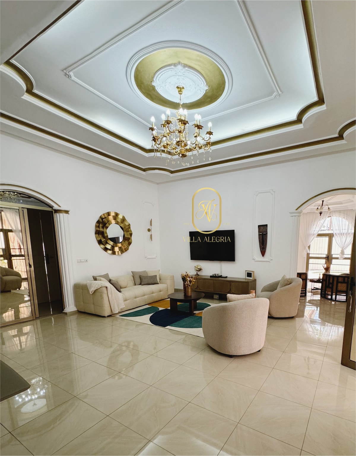 The living area features a high ceiling adorned with intricate moldings and a central chandelier. Neutral-colored sofas are arranged around a coffee table on a patterned rug. A mirror and decorative wall art accent the space, with natural light filtering through large windows with sheer drapes.