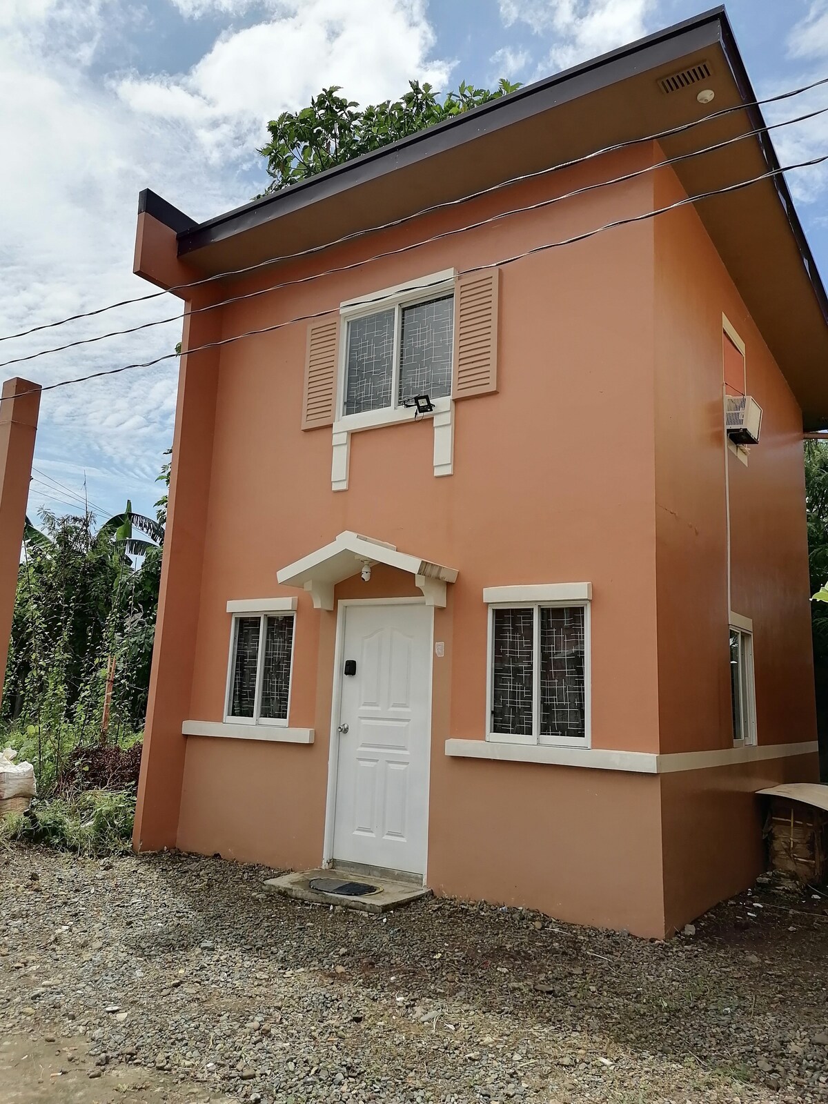 A newly constructed two-story house features a warm orange exterior with white trim. The front door is flanked by two windows, each adorned with shutters. Lush greenery is visible on the rooftop, while a gravel pathway leads to the entrance.