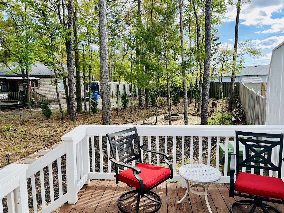 A wooden deck provides outdoor seating with two black chairs featuring red cushions, alongside a small round white table. Lush trees and greenery surround the area, creating a natural setting that offers a sense of tranquility.