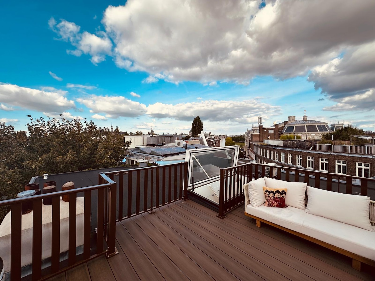 A spacious rooftop terrace is captured, showcasing panoramic views of Amsterdam's skyline under a partly cloudy sky. Comfortable outdoor seating is visible, surrounded by a railing with glass panels, allowing for unobstructed sightlines of the surrounding architecture and greenery.