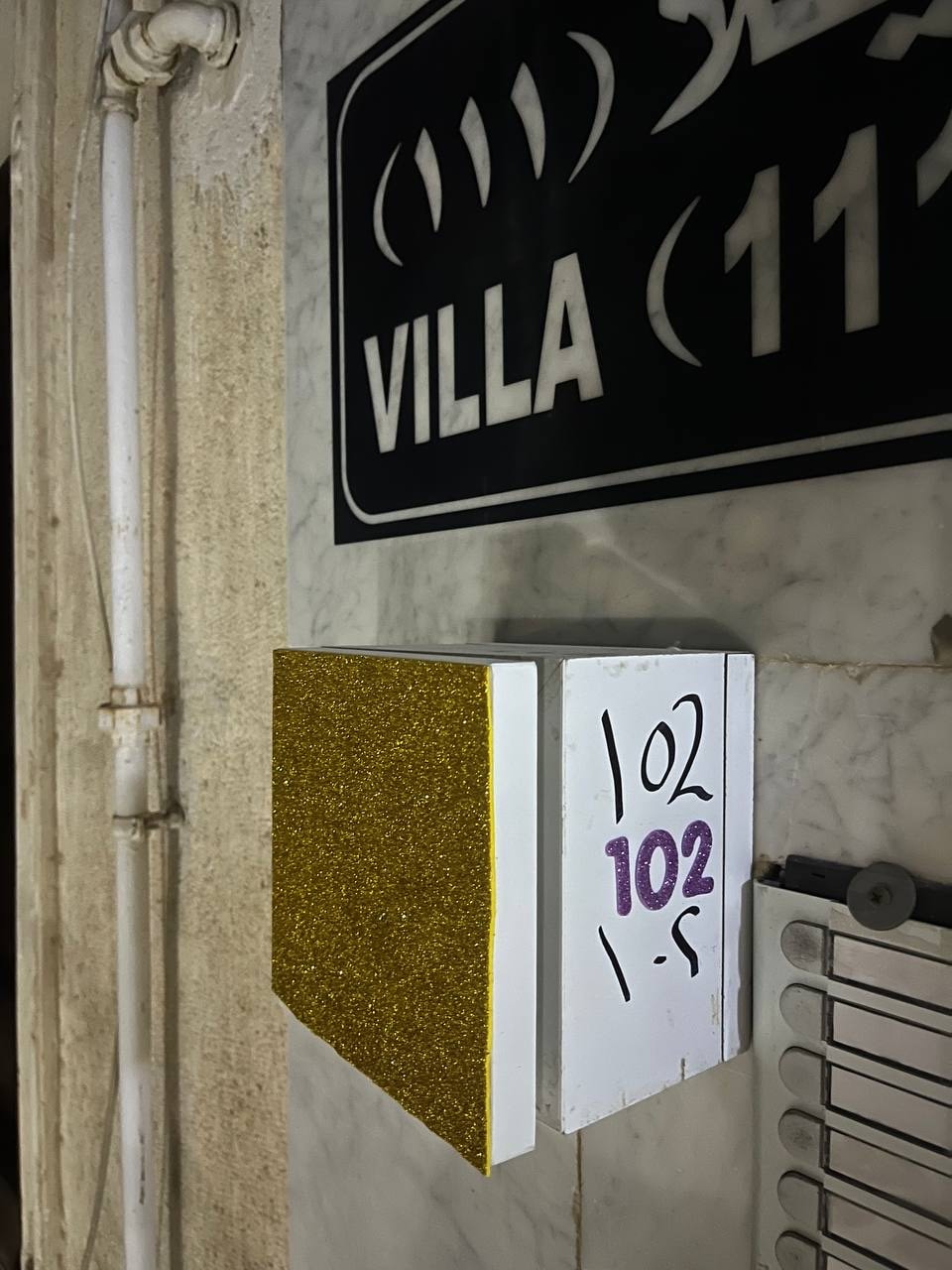 The image shows a close-up of the entrance to a villa, featuring a decorative sign labeled 'VILLA' above a mailbox. The mailbox displays the number '102' in bold, vibrant paint, surrounded by a gold glitter border. Key slots are visible below the mailbox.