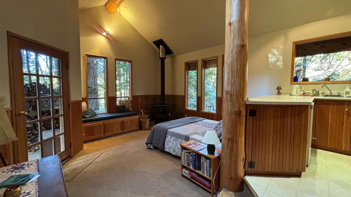 The open-plan living area features large windows that invite natural light and provide views of the surrounding cedar forest. A spacious bed is situated next to a small kitchen area, and a comfortable seating nook is visible near the window, creating a cozy retreat.