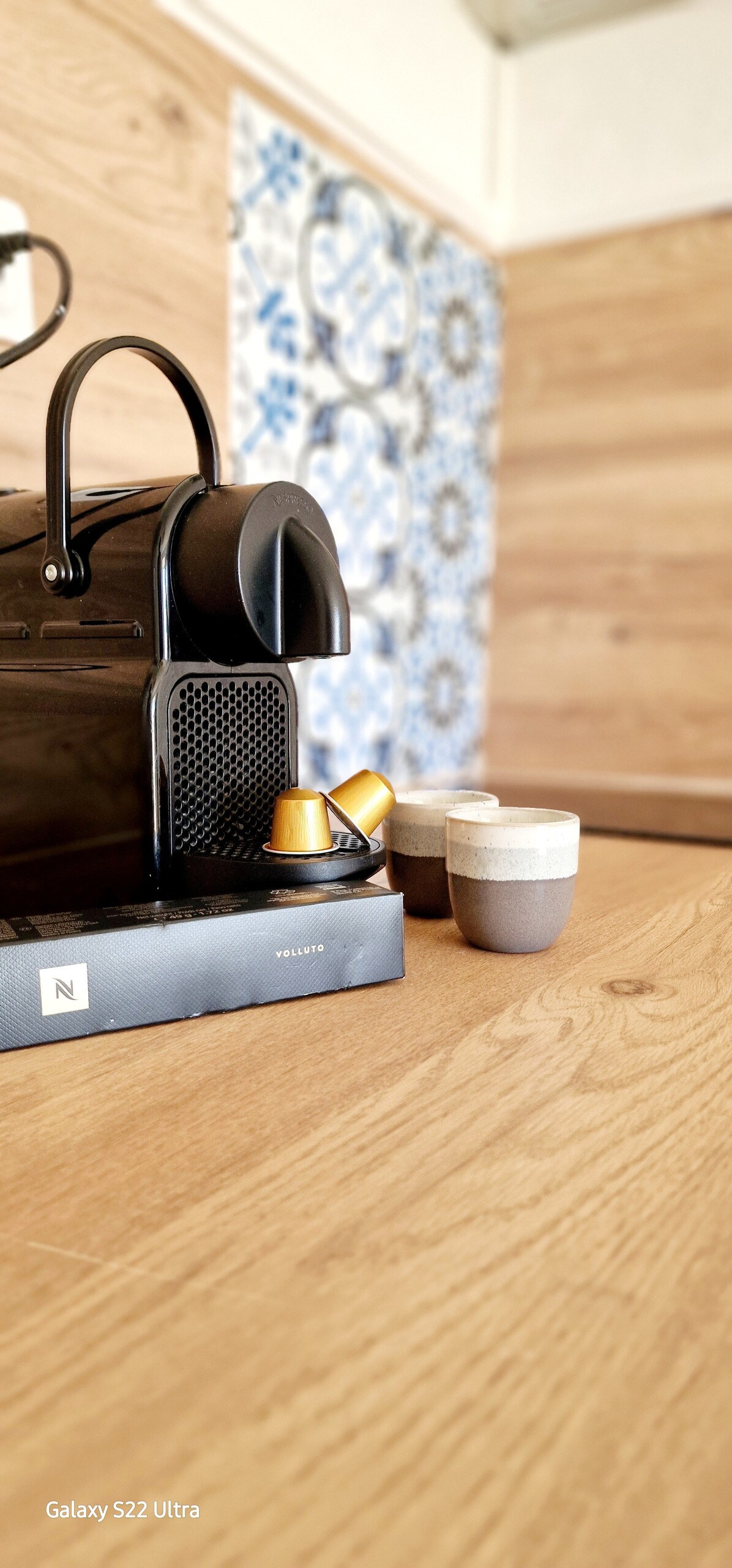 A Nespresso machine is positioned on a wooden countertop, accompanied by two ceramic cups in contrasting colors. The background features decorative tiles with intricate patterns, enhancing the space's design while providing a functional coffee area.