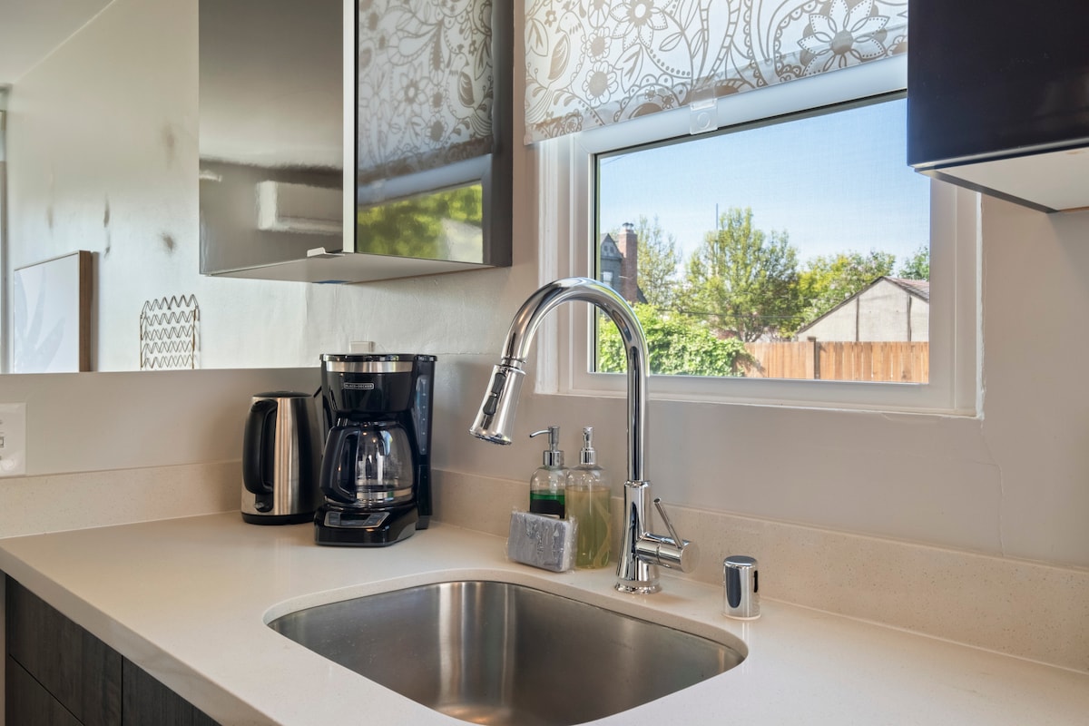 The kitchen area is equipped with a sleek countertop and a stainless steel sink. A coffee maker and kettle are positioned next to the window, allowing natural light to enhance the workspace. Soft fabric shades provide privacy while letting in light.
