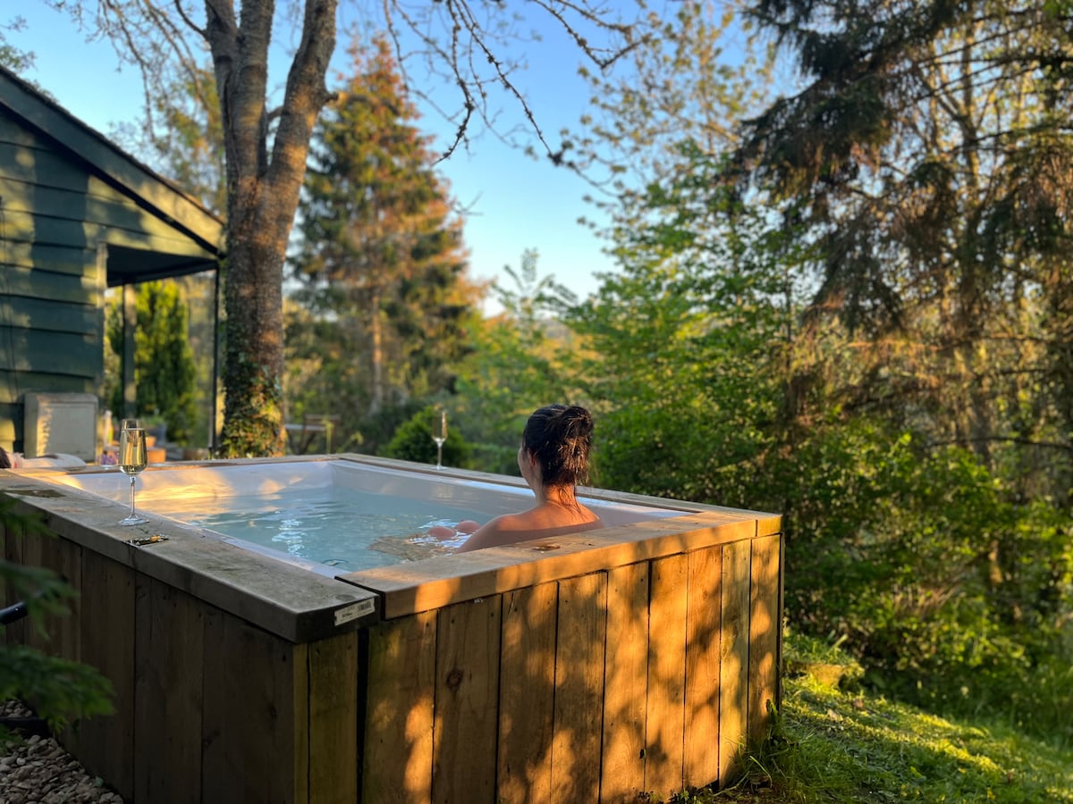 A wood-fired hot tub is situated in a serene outdoor setting, surrounded by lush greenery and trees. Sunlight filters through the branches, casting dappled shadows on the tub. The tranquil scene invites relaxation while offering views of the natural landscape.