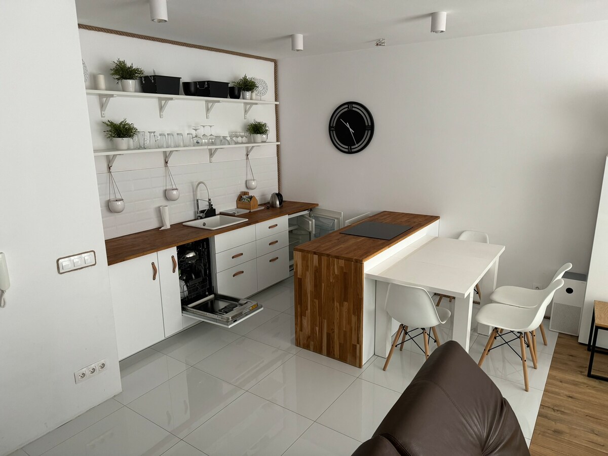 A modern kitchen area is presented with a minimalist design, featuring white cabinetry and a wooden countertop. A dining table is positioned nearby with four chairs. Clean lines and a bright atmosphere are enhanced by natural light reflecting off the glossy floor tiles.