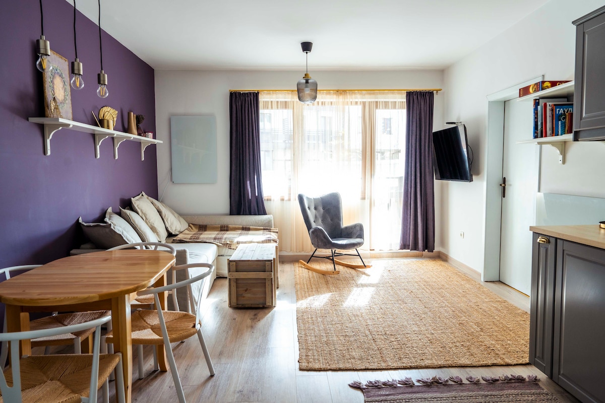 The living area features a cozy seating arrangement with a stylish armchair and a sofa, complemented by a round dining table. Natural light fills the space from the large window, which is adorned with sheer curtains, enhancing the warm tones of the purple accent wall.