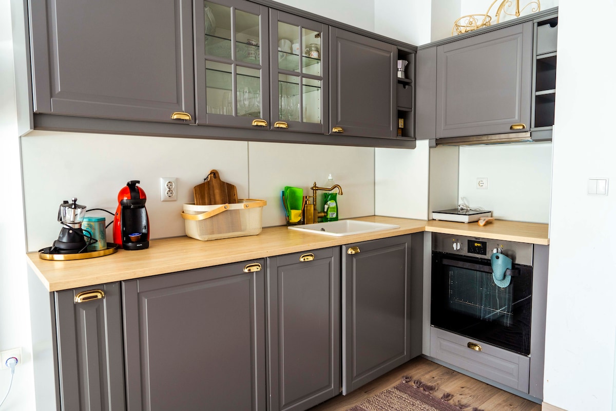 The kitchen features dark cabinetry with sleek hardware and a wooden countertop. Appliances include a kettle, coffee maker, and oven. A sink is positioned next to a large window, allowing natural light. Glassware and utensils are neatly arranged on the shelves.