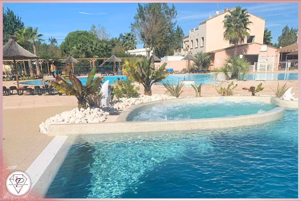 A spacious pool area features a blend of clear water and tropical landscaping, including palm trees and decorative plants. The design includes a children's area and palm-thatched shaded seating, creating a relaxing outdoor environment. Lounge chairs are positioned nearby for leisure.