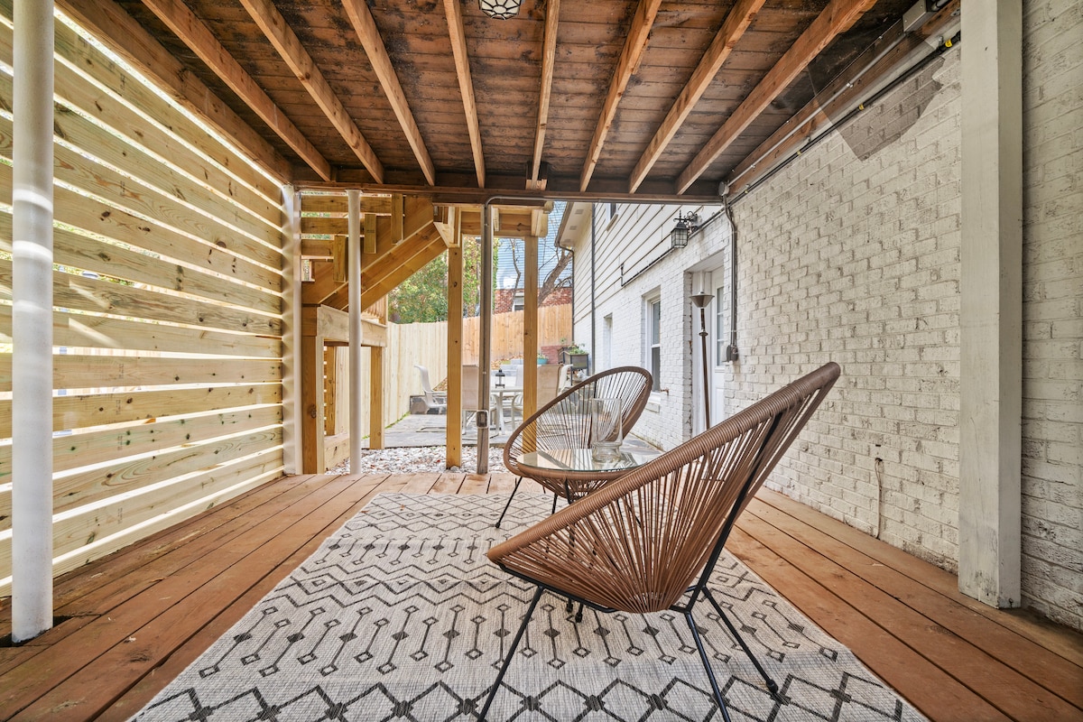 A lower deck area features two stylish woven chairs positioned around a smooth wooden table, all set on a decorative area rug. The space is framed by wooden beams and offers an open view of the yard.